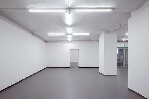 Das Foto zeigt einen leeren Ausstellungsraum mit reinweißen Wänden und Neon-Beleuchtung, im Hintergrund einen Eingangstüre aus Glase und ein Durchgang in einen anderen Raum