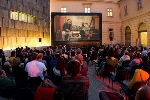 The photo shows an open-air cinema, a movie screen in the background, the audience sitting on chairs in front of it, some wearing headphones