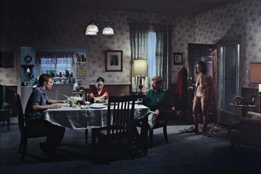 The picture by Gregory Crewsdon shows a room in a gloomy mood, three people sitting at a dining table, a naked, depressed-looking woman comes in at the door