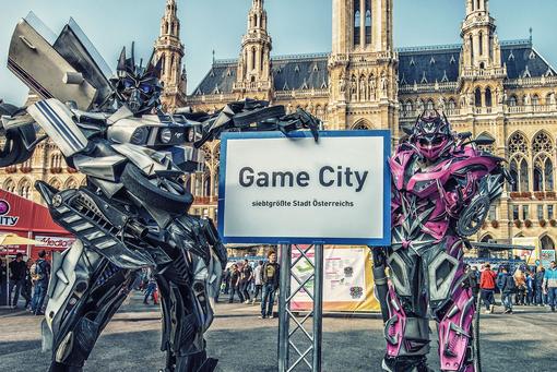 Eine Fotomontage mit zwei Transformer-Figuren, die die Ortstafel Game City halten, im Hintergrund das Wiener Rathaus und Besucher:innen auf dem Wiener Rathausplatz