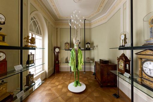 Gezeigt wird ein Biedermeier-Raum mit unterschiedlichen historischen Uhren, in der Mitte steht ein Modell in Lindgrün von der Modedesignerin Florentina Leitner