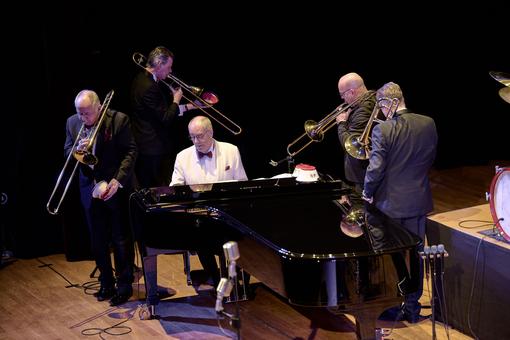 The photo shows a pianist in a white suit at a grand piano, around which four trombonists walk and play music