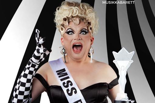 The photo shows drag queen Grazia Patricia with her blonde hair up, black cocktail dress, white and black checkered long gloves and holding an award in her hand