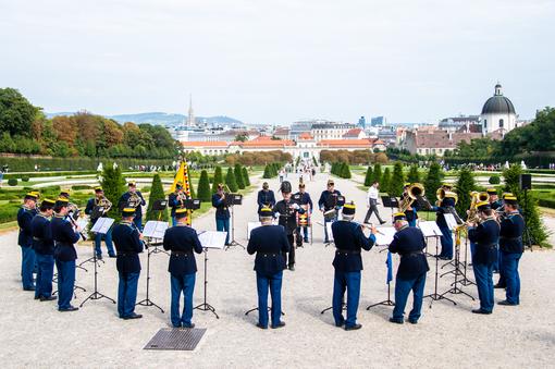 Das Foto zeigt die traditionelle Blasmusik-Kapelle der Original Hoch- und Deutschmeister im Schlosspark Belvedere vor dem Oberen Belvedere mit Blick auf den Park, auf das Untere Belvedere. Die Musiker tragen traditionelle dunkelblaue Uniformen.
