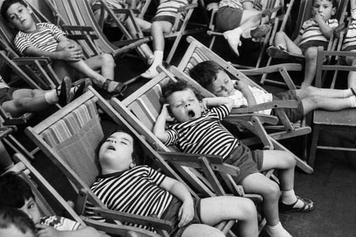 Das Schwarzweiß-Foto zeigt kleine Kinder in gestreiften T-Shirts und kurzen Hosen, die in gestreiften Liegestühlen liegen bzw. schlafen