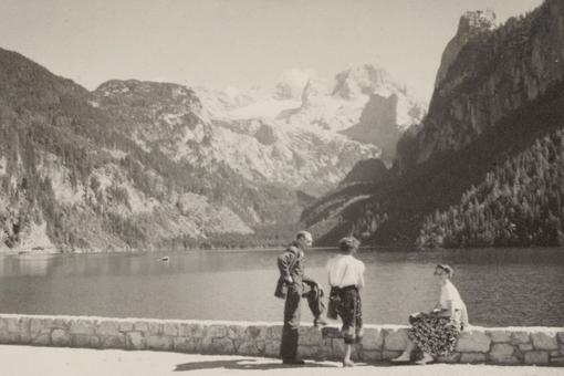 A classic vacation photo in black and white: a mountain range and forest in the background, a lake in front of it. Two women and a man on the shore, looking at the lake and into the camera respectively