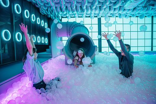The photo shows a woman and a man having fun in a ball pool, a girl slips out of a tube into this ball pool, the scene is bathed in blue and magenta light at the edges