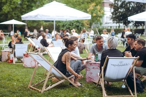 Das Foto zeigt eine Szene in einem Park: Menschen sitzen in Liegestühlen, unter Sonnenschirmen und genießen Drinks