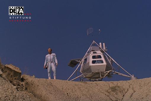 The photo shows a futuristic silver space capsule in the 1960s, next to it a man in a space suit, both standing on rocky ground, behind them the deep blue sky