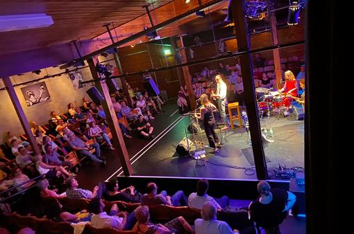 The photo shows a concert situation in the Theater am Spittelberg, in the middle the stage with the musicians: Musician on drums, three guitar players, the audience is seated on three sides of the stage