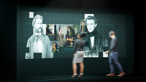 Zu sehen ist eine Ausstellungsansicht des Johann Strauss Museum – New Dimensions, eine multimediale Wand mit Bildern von Johann und Josef Strauss und Ausschnitten aus Filmen über die Strauss-Familien, davor zwei Besucher:innen des Museums