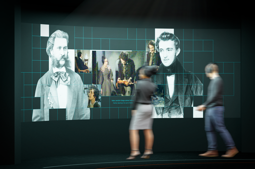 Zu sehen ist eine Ausstellungsansicht des Johann Strauss Museum – New Dimensions, eine multimediale Wand mit Bildern von Johann und Josef Strauss und Ausschnitten aus Filmen über die Strauss-Familien, davor zwei Besucher:innen des Museums