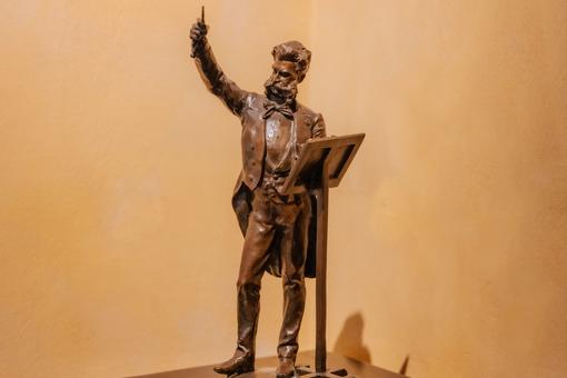 The photo shows a brown statuette of the composer Johann Strauss. Johann Strauss stands in a Frank at a lectern and conducts