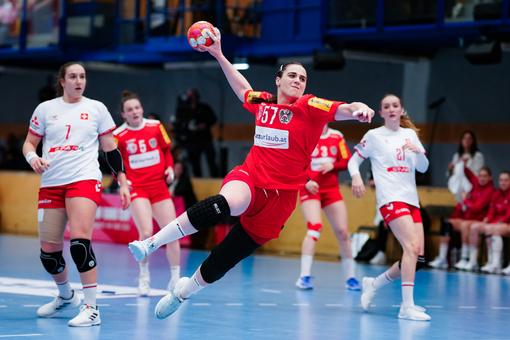 Das Foto zeigt eine Szene aus einem Handballspiel der Damen, im Vordergrund eine Spielerin im roten Trikot, die mit dem Ball in der Hand zum Schuß ausholt