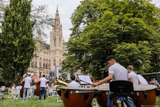 The photo shows the Vienna City Hall Park with young musicians and their instruments on white park chairs, in the background the Vienna City Hall