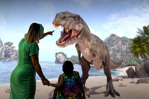 Ausstellungsansicht: immersiver Tyrannosaurus Rex in einer Insellandschaft, davor eine Besucherin mit zwei Kindern in ihrer Begleitung