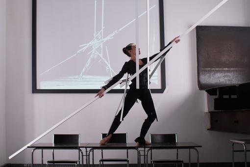 The photograph shows a woman in black clothing standing on a modern table. She is holding white, long slats in the diagonal of her outstretched arms and two in the vertical of her body