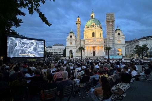 Das Foto zeigt ein Open-Air-Kino, eine Kinoleinwand mit Publikum davor, im Hintergrund die Karlskirche