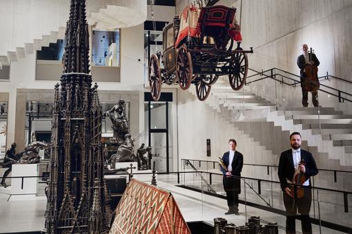 The photo shows musicians from the Vienna Symphony Orchestra with their instruments standing between the exhibits in the large hall of the Wien Museum