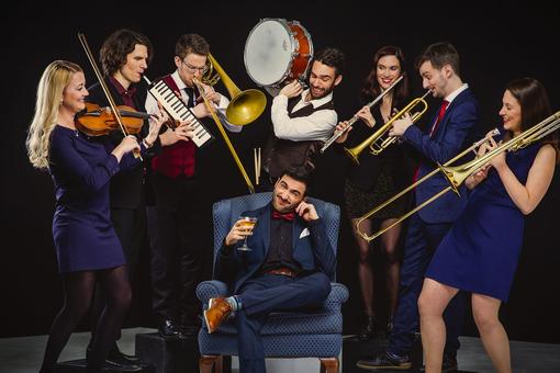 You can see the klezmer band Octopus, three women and three men standing with their instruments around a blue chair, on which another band member is sitting with a glass in his hand and covering his ear