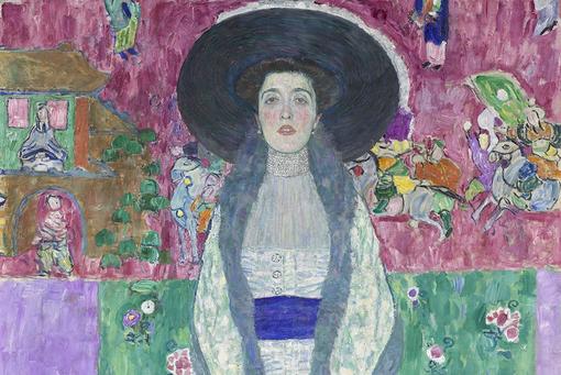 Gemälde von Gustav Klimt, das Adele Bloch-Bauer mit großem Hut und bodenlangem Kleid zeigt. Im Hintergrund bunte Blumenornamentik und Bilder einer chinesischen Tapete.  