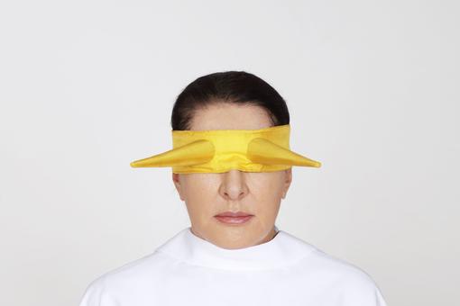 Photo of artist Marina Abramović in a pure white top against a white background. She wears a yellow silk blindfold with two outstanding tips in place of the eyes.
