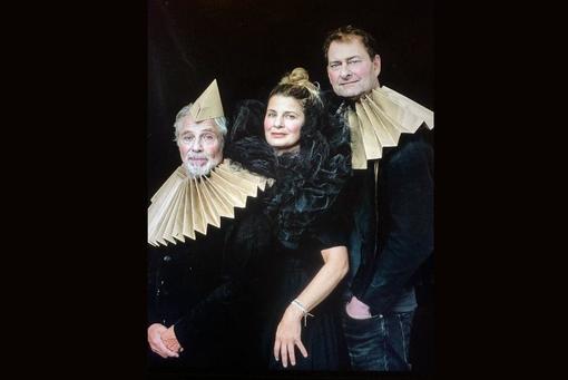 The photo shows the three main actors in black costumes, beige and black, large collars