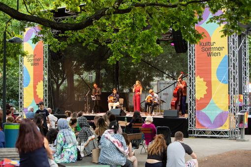 Photo of an open air situation in a park: a band playing music on a stage under trees, in front of it the audience