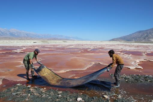 Two men face each other in a copper-colored inhospitable landscape and hold a photographic paper about three meters long in their hands