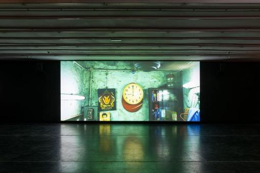 Exhibition view showing a projected large-scale image. In the center a clock, next to it a display case and pictures, the scene is immersed in green