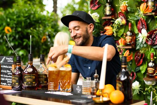 Photo of young bartender with dark cap, in front of him different drinks on a counter