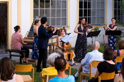 The photo shows a musical ensemble with piano, flutes, violin and cello open air in a green courtyard, with an audience in the foreground 