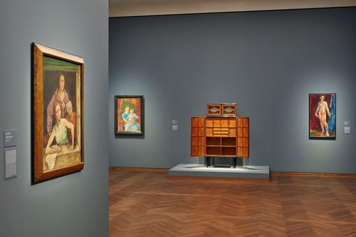 Exhibition view: three paintings and a marquetry cabinet in a room with gray-blue walls and a herringbone parquet floor