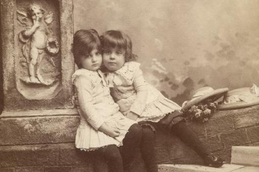 Schwarzweiß-Fotografie der Schwestern Alma und Margarete Schindler, zwei kleine Mädchen in weißen Kleidern, aneinander geschmiegt, an eine Stele mit einem Engel gelehnt