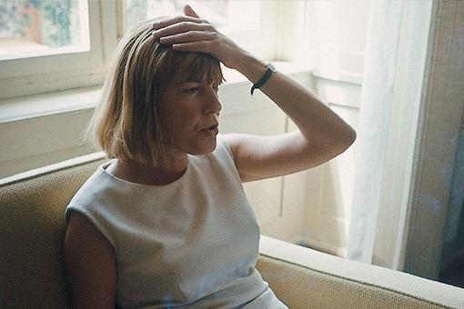 Foto von Ingeborg Bachmann, die in einem weißen Kleid auf einer Couch sitzt und sich mit der linken Hand auf den Kopf greift