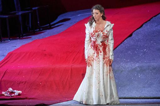 Szenefoto aus der Oper "Lucia di Lammermoor": zu sehen ist die Hauptdarstellerin in einem mit Blut beschmierten, weißen, langen Kleid, die fassungslos in Richtung Publikum blickt, im Hintergrund rote und blaue Stoffbahnen
