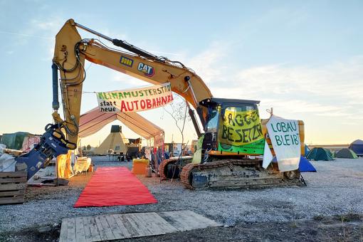 The photo shows part of the Lobau-bleibt! in Vienna. In the foreground a large yellow excavator covered with protest banners and posters, behind it tents and a wooden pyramid