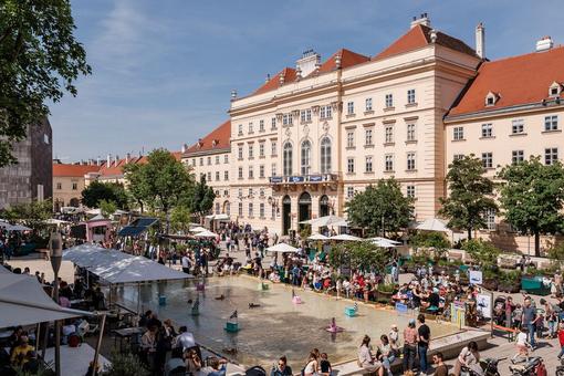 You can see the main courtyard of the MuseumsQuartier Wien in summer with lots of people and plants, in the middle a rectangular water basin