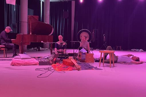 Photo of a stage scene: three men and a woman playing music sitting and lying, around the persons numerous instruments, the stage in magenta light