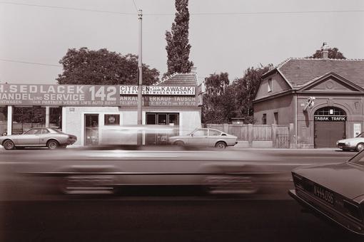 Das Schwarzweiß-Foto zeigt einen Teil der vielbefahrenen Triester Straße in Wien 1982-83, im Hintergrund das Gebäude eines Autohändlers und eine Trafik, im Vordergrund die fahrenden Autos