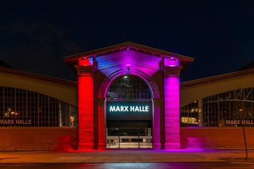 Foto des Haupteinganges der Marx Halle bei Nacht, in rotpinkes Licht getaucht. Das Portal hat zentral ein oben rundes Tor, mit seitlich zwei Säulen, darüber ein Giebeldach. Über dem Eingang der Schriftzug Marx Halle