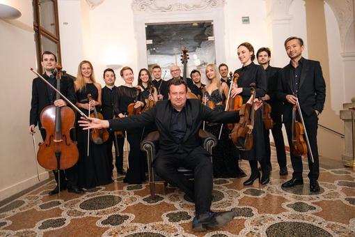 The photo shows the Morphing Chamber Orchestra, the musicians are dressed in black, holding their instruments in their hands. The director of the orchestra sits in a leather chair with his arms outstretched in front of the musicians