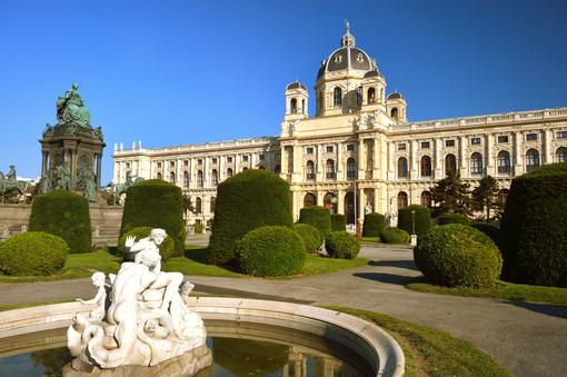 Photo from the Natural History Museum Vienna, in the foreground a round fountain with sea figures in the middle, a well-kept park with accurately cut bushes, left the monument of Empress Maria Theresia