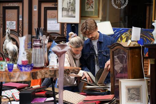 Szene auf einem Flohmarkt: eine Frau und ein Mann sind in alte Alben vertieft, rund um sie befinden sich alte Bücher, Fotoalben, Bilder, Glasgegenstände etc.