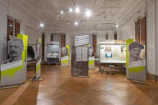Exhibition view: different display panels in a historicist room with white stucco ceiling and parquet floor