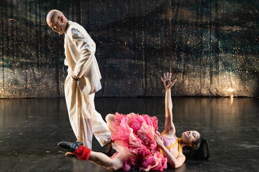 Photo of a performance: a dancer in pink puffy tulle skirt lying on the floor with her right arm up and a man in a white suit turning away