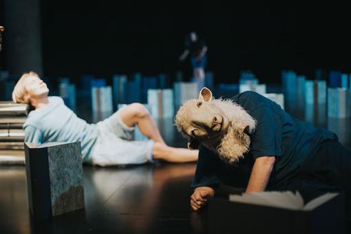 Scene with two people on stage between books, one of the people is carrying a sheep's head