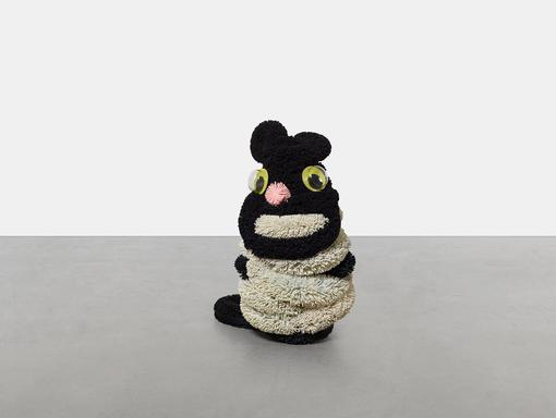 The photo shows a sculpture created by Damien Hirst of a sitting cat made of fluffy black and beige material with large green-yellow plactic eyes