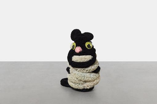Das Foto zeigt eine von Damien Hirst geschaffene Skulptur einer sitzenden Katze aus flauschigem schwarzem und beigen Material mit großen grüngelben Plaktikaugen