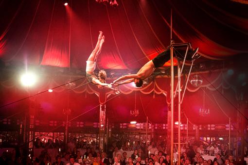 Photo of a trapeze acrobatics in the palace of mirrors of the Palazzo, in the background the audience, the scene is illuminated red